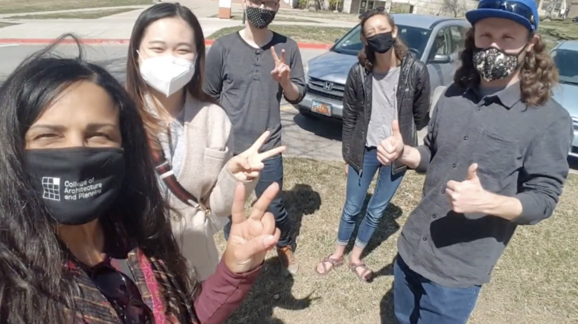 Five university students are wearing masks and giving approval hand signs (thumbs up, Vee). They are standing on grass. Two cars and a building are visible behind them.