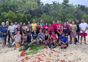 A large group of about 30 people pose for a photo on the beach. There are red flowers and huge leaves on the sand in front of them.
